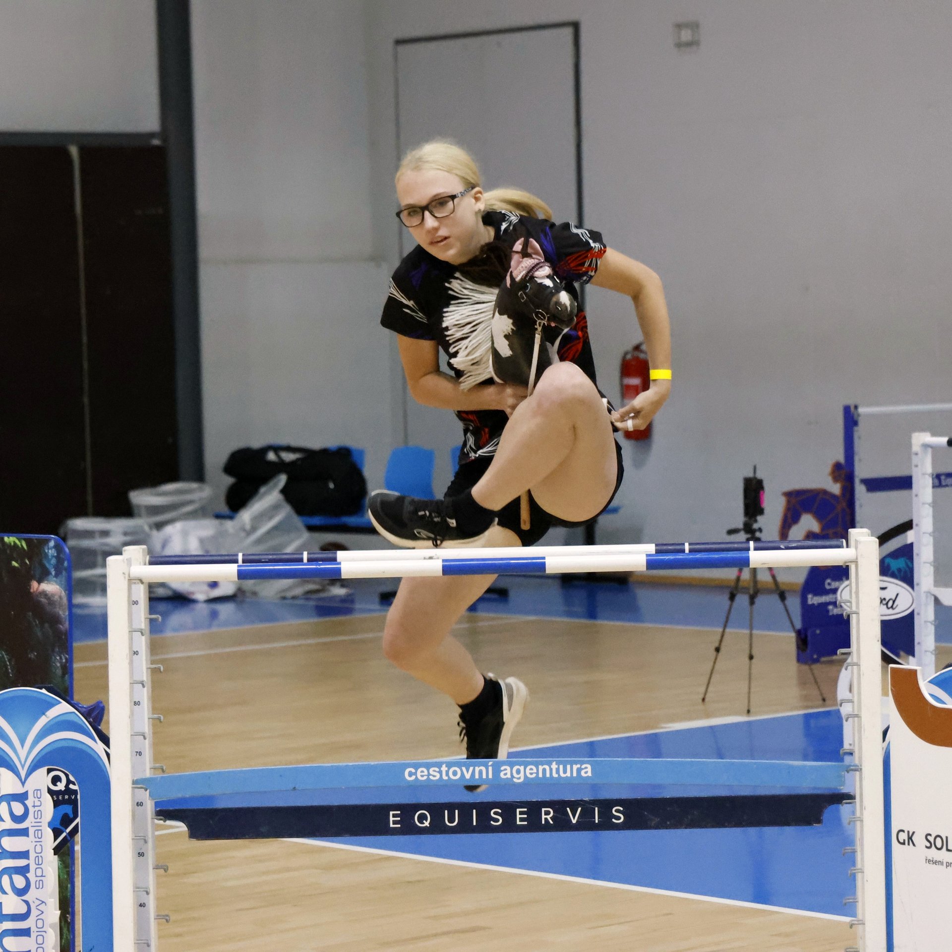 Závodnice ve skoku mohutnosti přeskakuje vysokou laťku s Hobby Horse koněm v ruce ve sportovní hale na mistrovství Evropy.